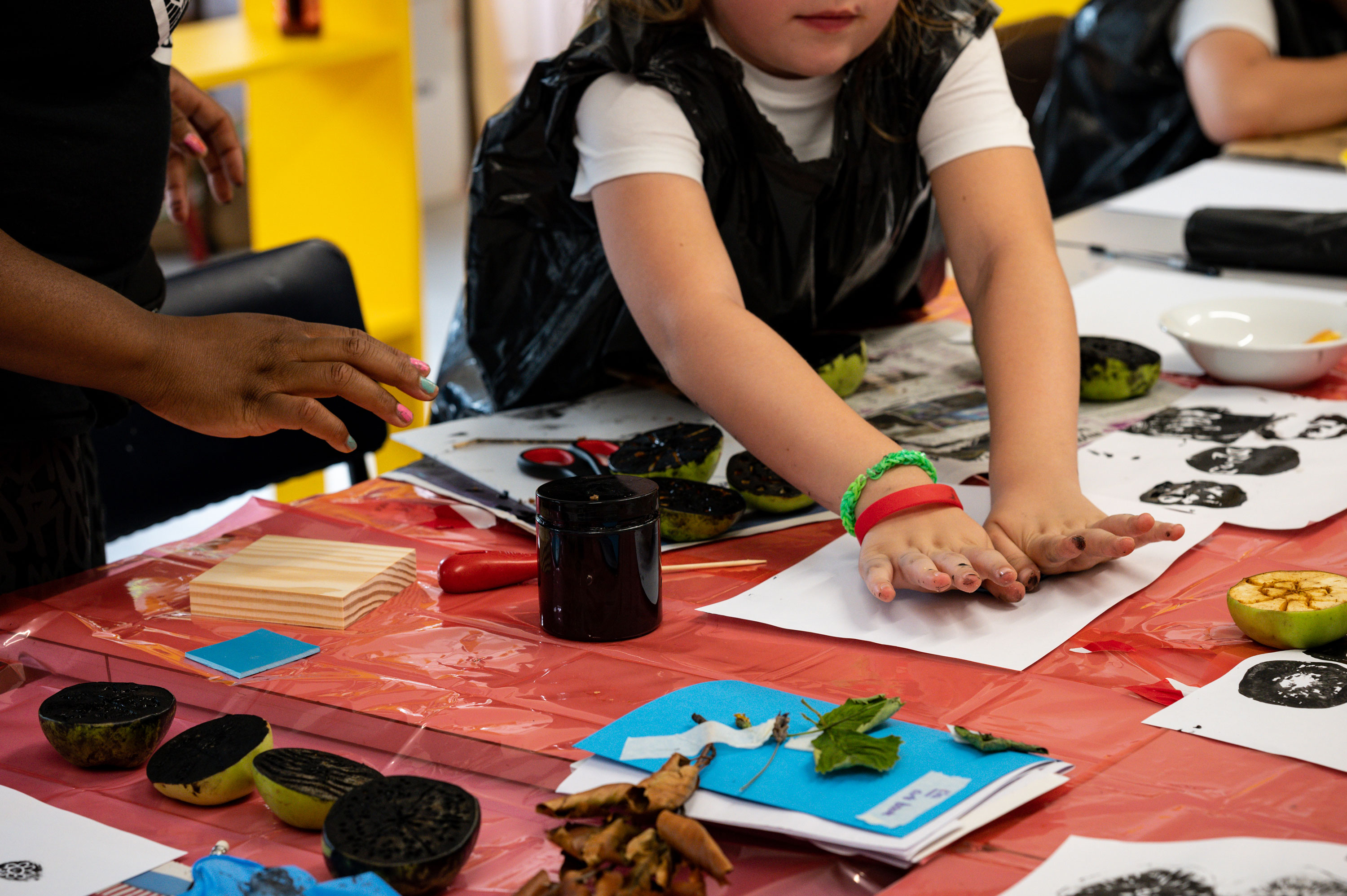 A table with paper, ink and leaves. A young girl using both hands to press down on a sheet of paper to make a print.