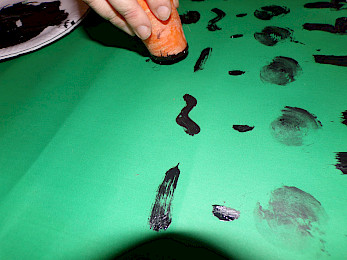 A hand using a carrot to create a pattern of black spots on a green backgroun