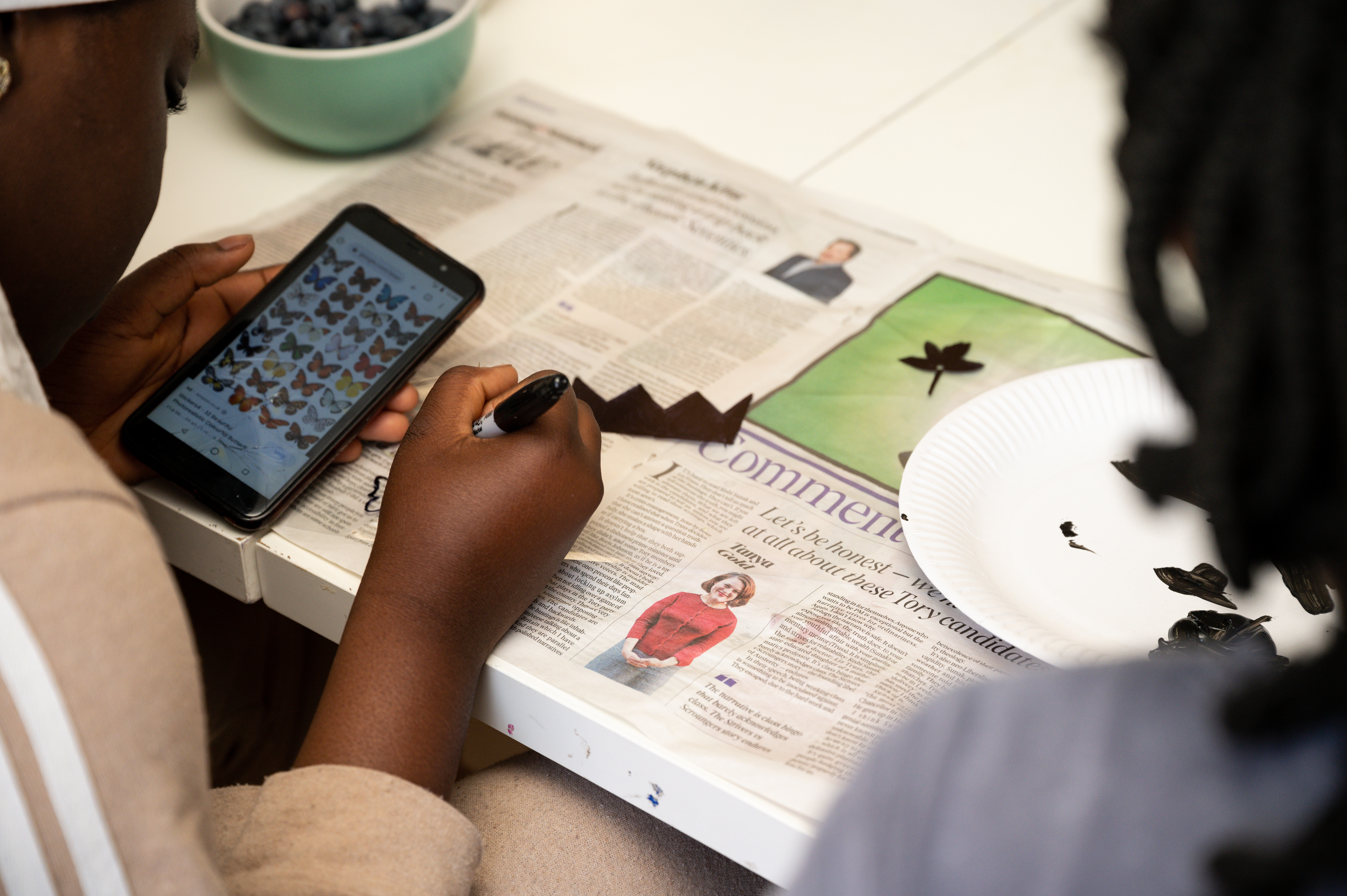 2 children sitting facing away from camera looking at butterful images on a phone and drawing with black pen and black paint