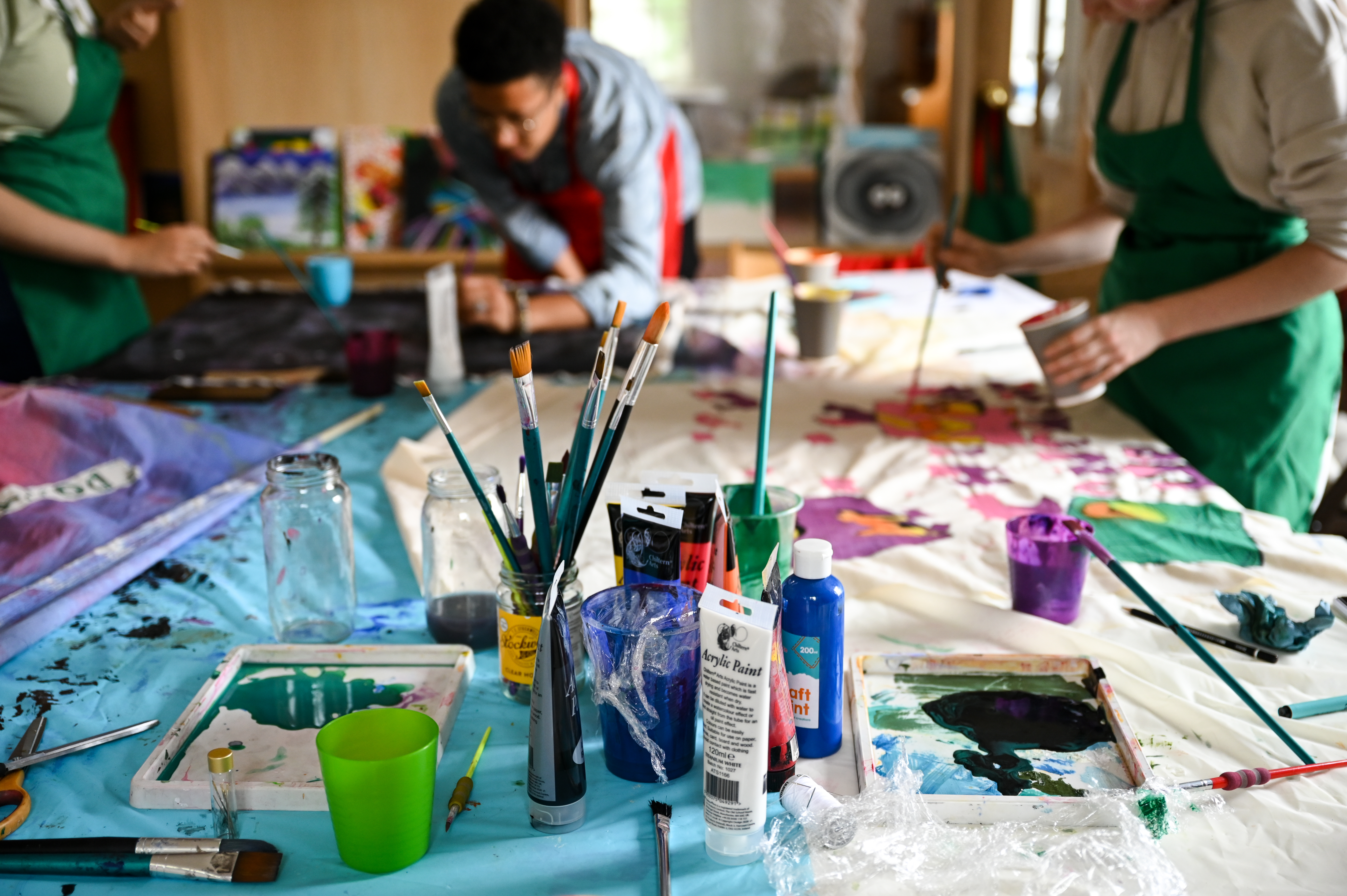 3 young people painting on messy table with lots of artworks spread out