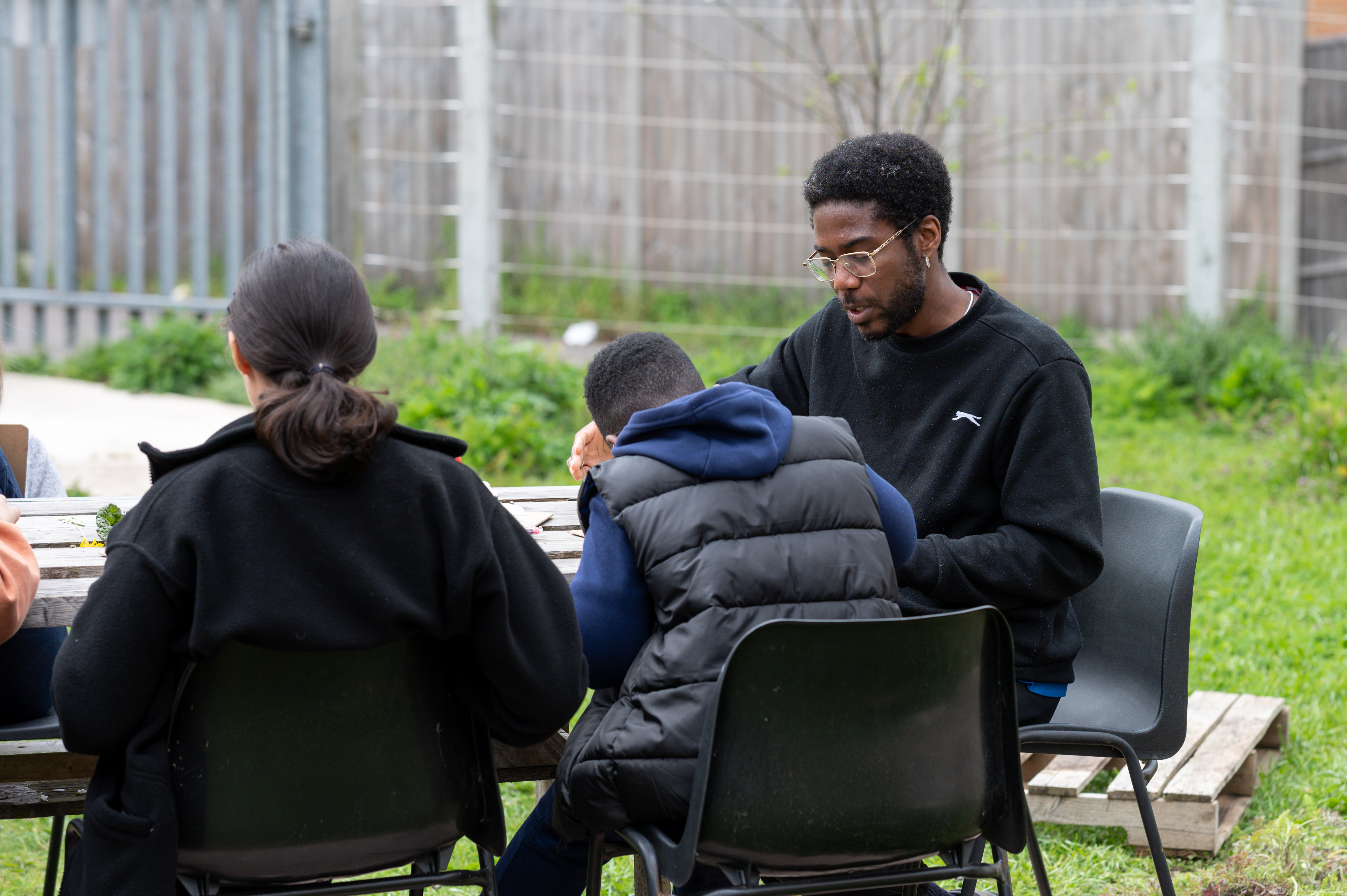 adult and 2 children sit at outside table in coats engaged in focused activity