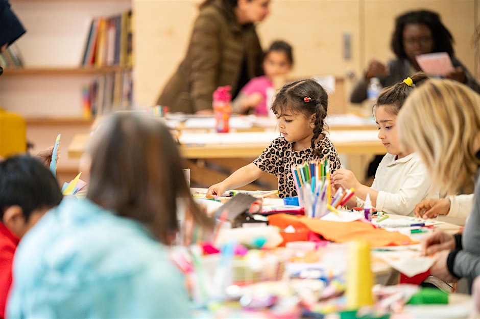 A photograph taken at the Women's Museum of a creative workshop. Children and parents are seated round tables making crafts, with pens, paper and paints on the table.