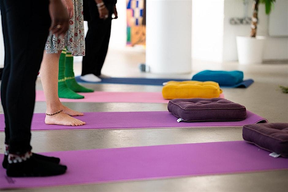 A photograph of four people standing at the end of their yoga mats, which are blue, pink and purple. They have cushions at the top of their mats.